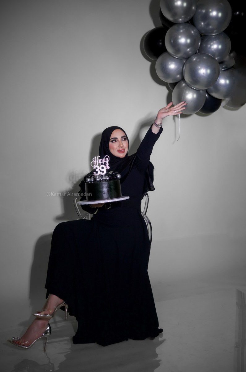 Woman in a black hijab holding a birthday cake beside black balloons, photographed by Kamar photography